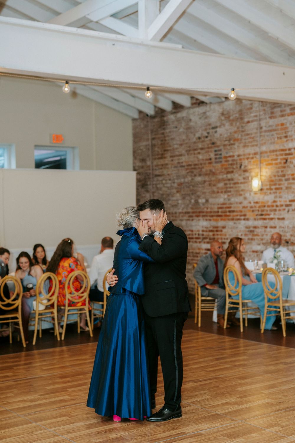 A groom in a black suit shares a first dance with his mother in a blue gown at a wedding reception with brick walls.