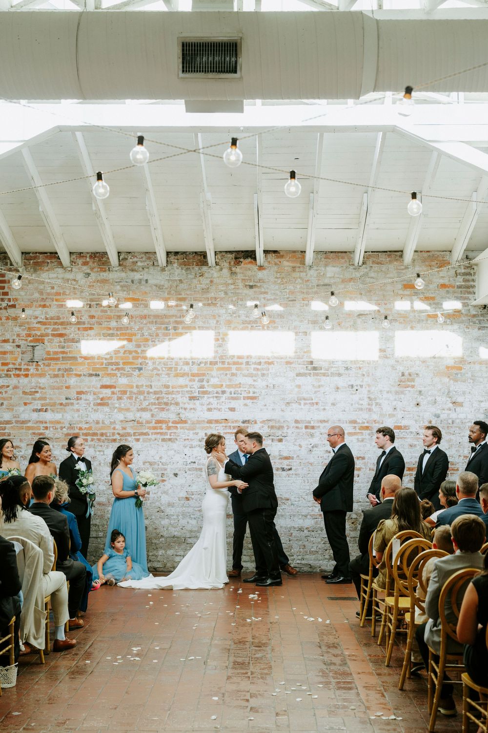 Indoor wedding ceremony with couple exchanging vows before brick wall, string lights overhead, guests seated in wooden chairs.