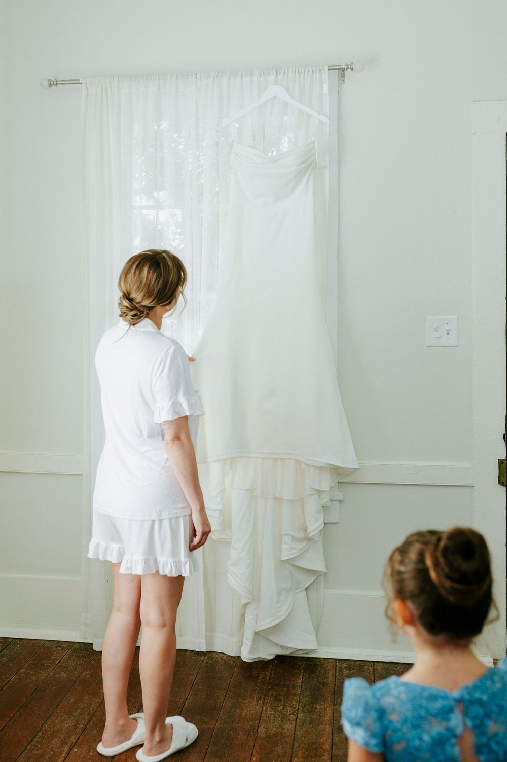 Bride in white robe admires her wedding gown hanging by a sunlit window, with a young girl watching nearby.