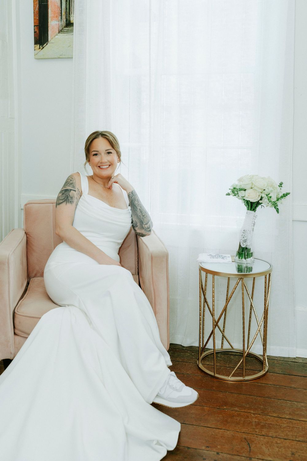 Smiling bride in white gown and sneakers seated in pink chair beside gold side table with white floral bouquet.