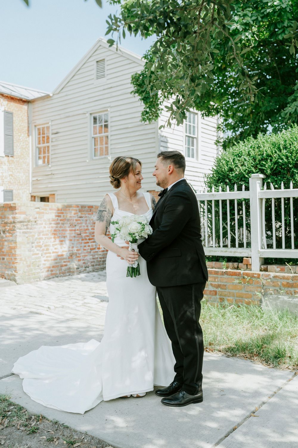 A bride and groom share a romantic moment outdoors, with the bride holding a white bouquet near a brick wall and white fence.