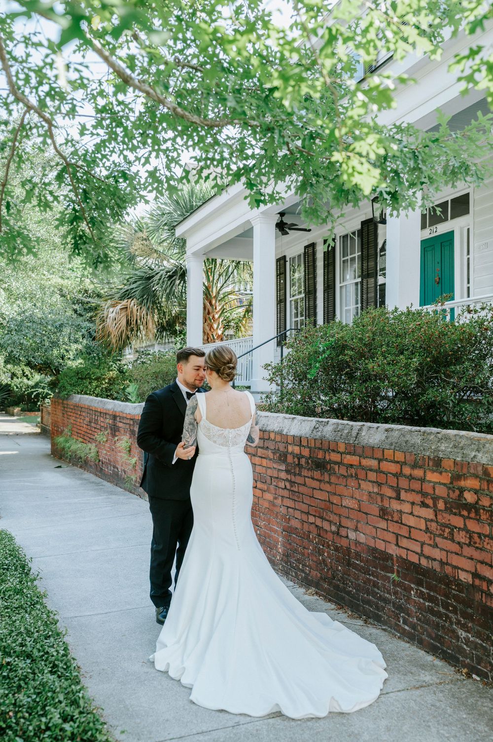 Bride and groom share a romantic moment on a brick sidewalk outside a historic Southern home surrounded by lush greenery.