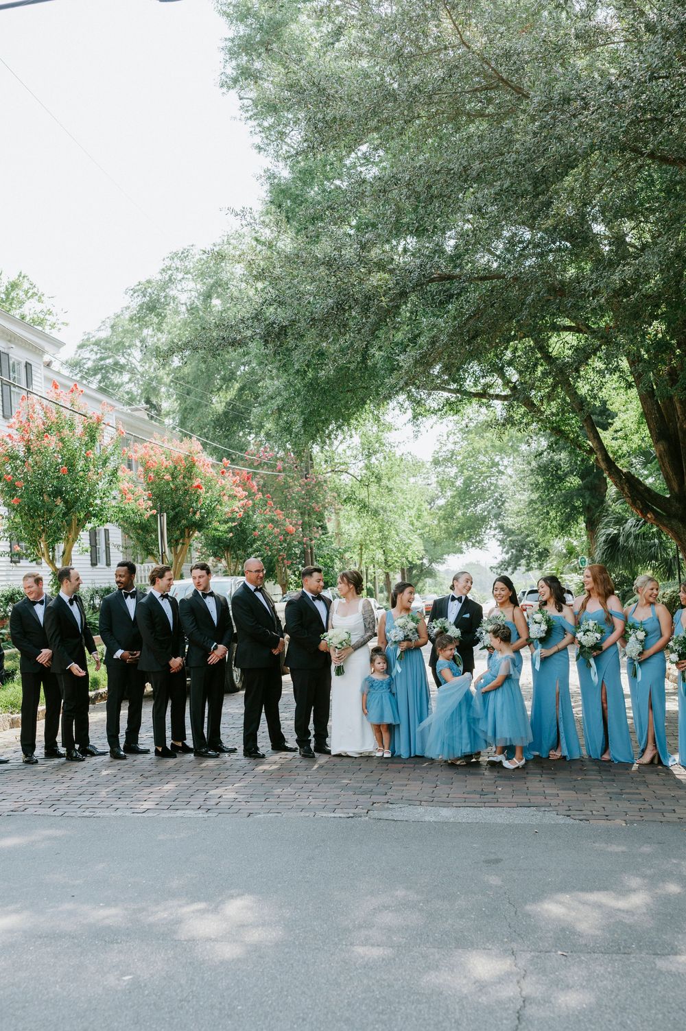 Wedding party poses outdoors, groomsmen in black tuxedos and bridesmaids in blue gowns, with bride and groom centered under lush trees.