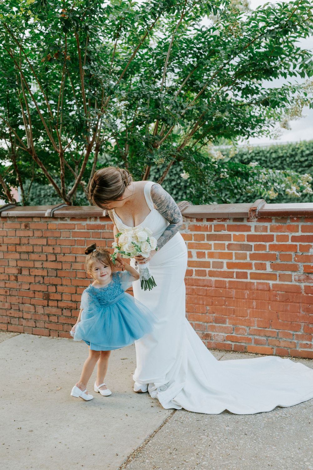 Bride in white gown bends to interact with flower girl in blue dress, holding bouquet near brick wall.