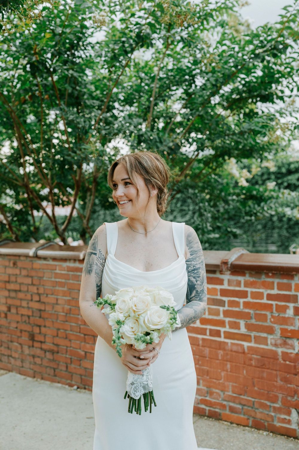 Smiling bride in white gown holds white rose bouquet, showcasing sleeve tattoos near a brick wall.