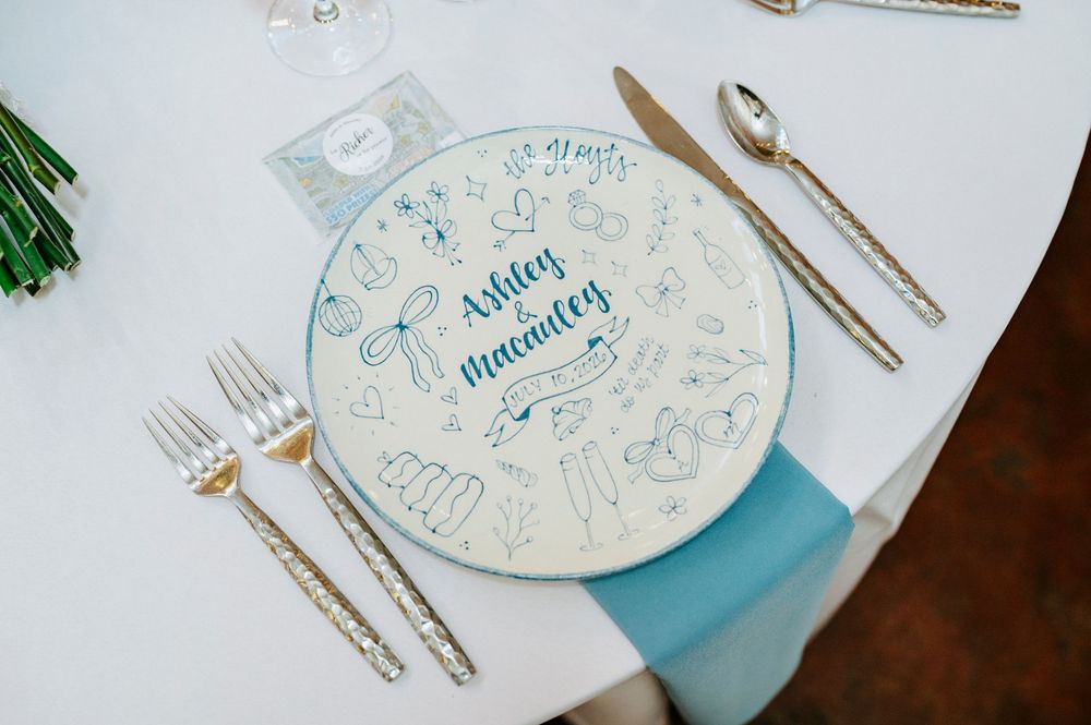 Custom illustrated wedding charger plate with blue hand-lettered names Ashley and Macauley, gold cutlery on white tablecloth.