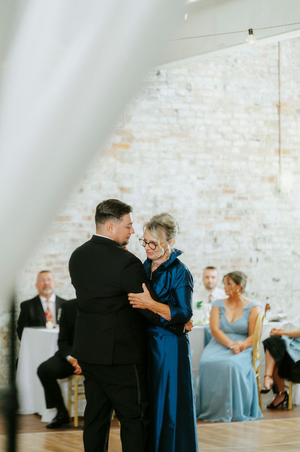 Groom shares a heartfelt mother-son dance at wedding reception in elegant venue with exposed brick walls.
