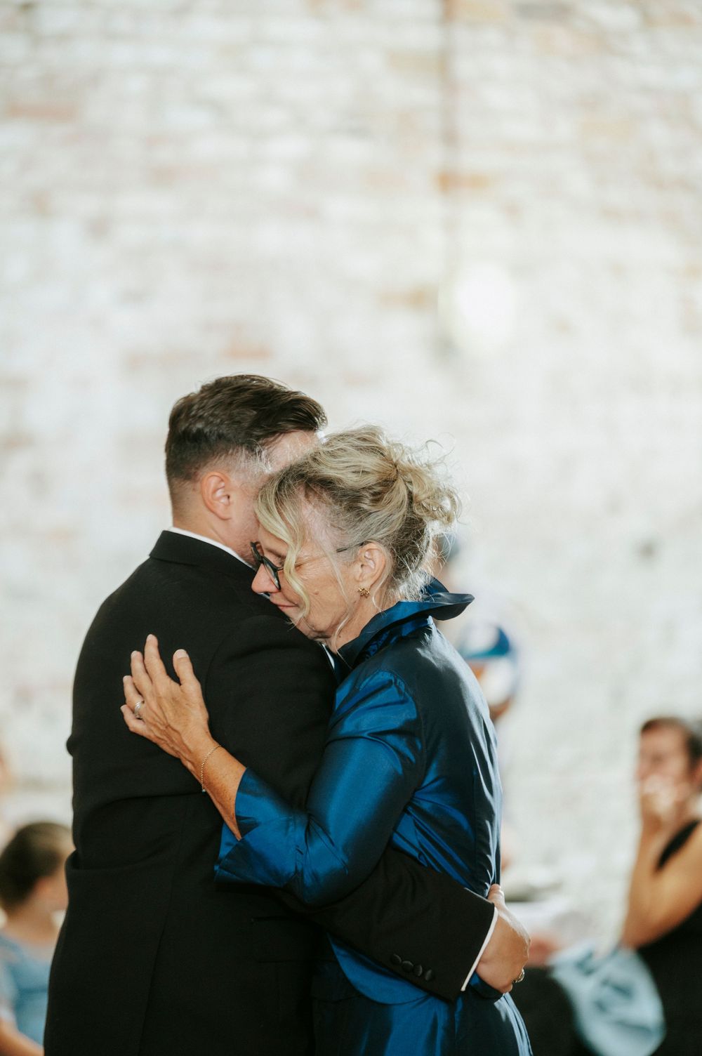 A groom in a black suit shares an emotional dance with an older woman in a blue dress at a wedding reception.