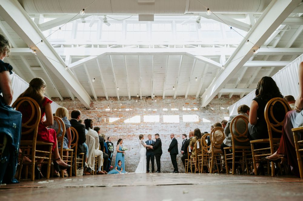Indoor wedding ceremony with guests seated in wooden chairs facing couple at altar under skylight roof.