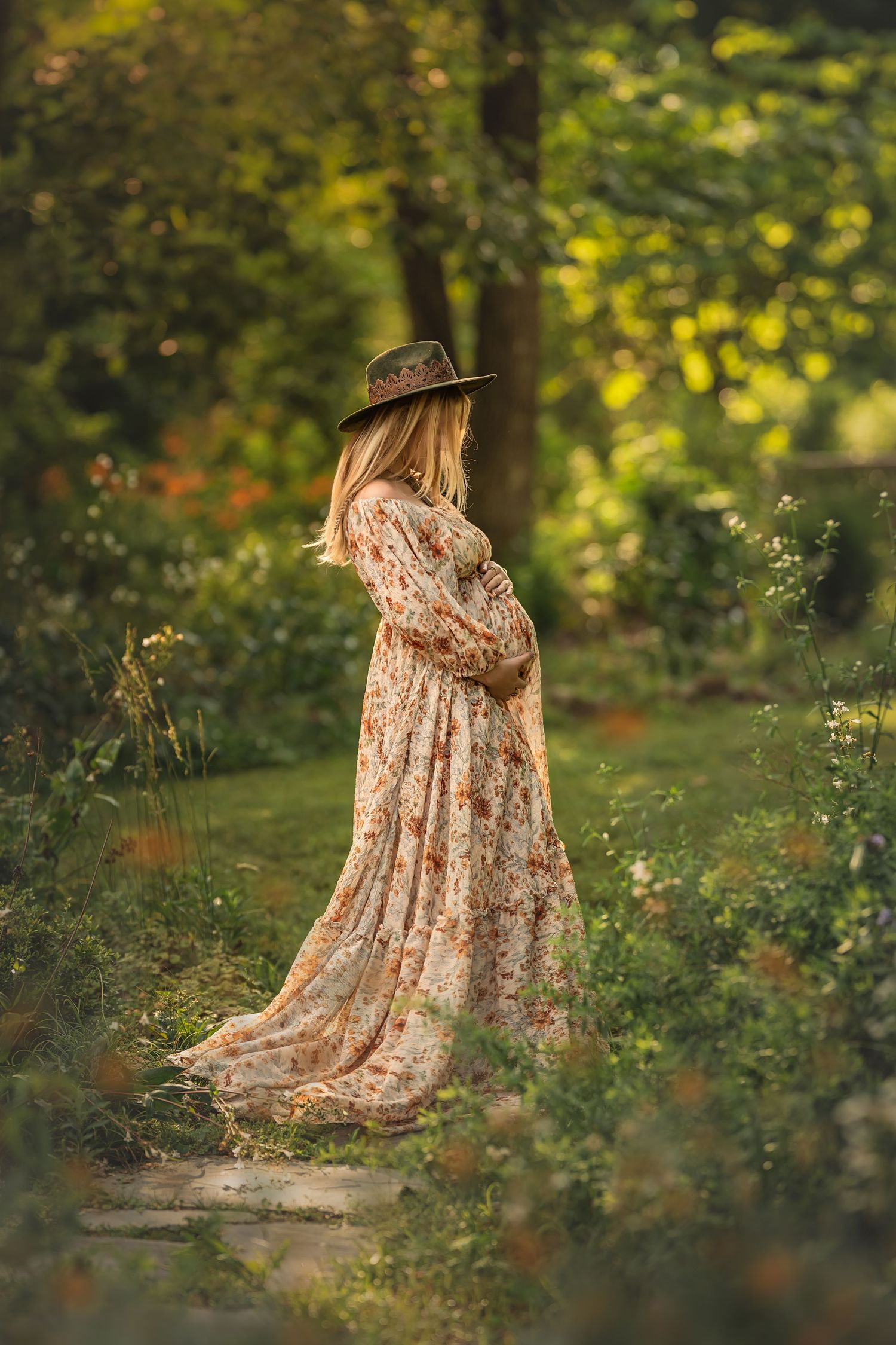 A pregnant woman in a floral maxi dress and brown hat stands in a dreamy garden setting with golden sunlit foliage.