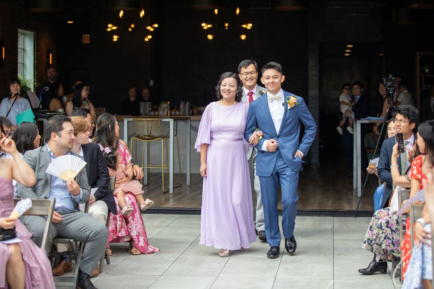 Groom in blue suit walks down wedding aisle with mother in lavender gown, guests seated on both sides.