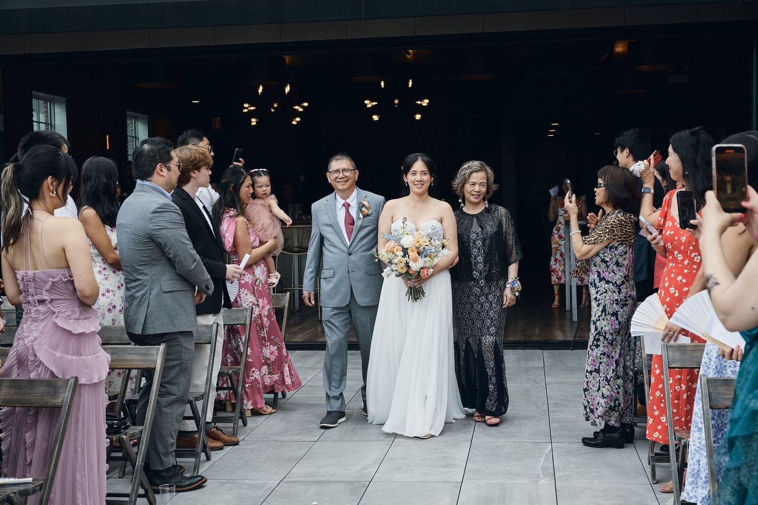 Bride walks down the aisle with parents at an outdoor wedding ceremony, guests watching and photographing.
