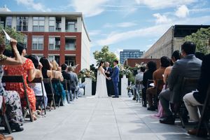 Outdoor wedding ceremony on urban rooftop with couple at altar, guests seated along aisle, city buildings in background.