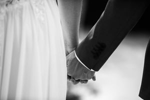 Black and white photo of a bride and groom holding hands, showing a tattoo on the groom's wrist.