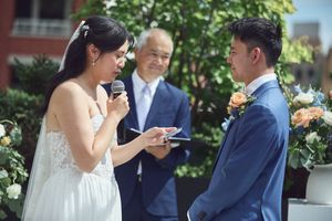 Bride reads wedding vows into microphone while groom listens during outdoor ceremony with officiant.