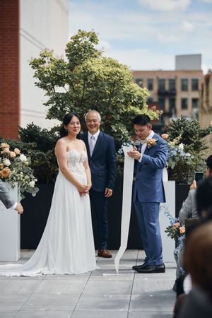Couple exchanges vows at rooftop wedding ceremony with officiant, floral arrangements, and city buildings in background.
