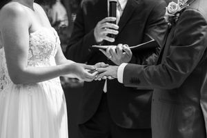Black and white photo of bride and groom exchanging rings during wedding ceremony with officiant in background.