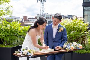 Bride and groom signing wedding register on rooftop venue surrounded by lush greenery and blue floral arrangements.