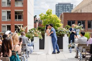 Couple shares first kiss at outdoor rooftop wedding ceremony surrounded by floral arrangements and seated guests in urban setting.