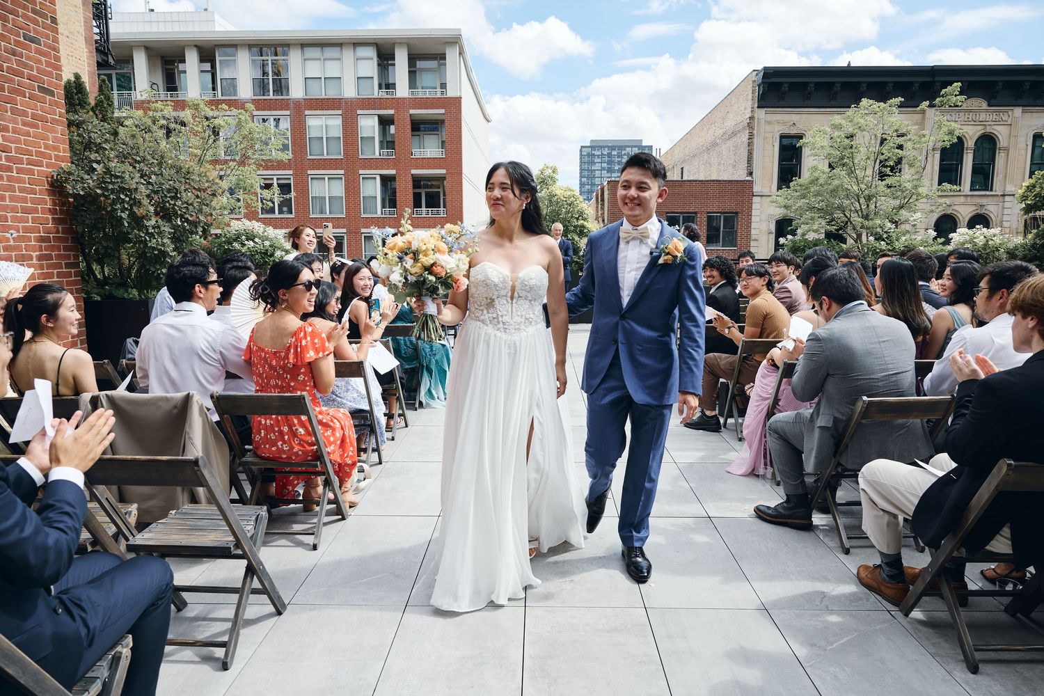 Bride and groom walk down outdoor rooftop wedding aisle, surrounded by seated guests, with urban buildings in the background.