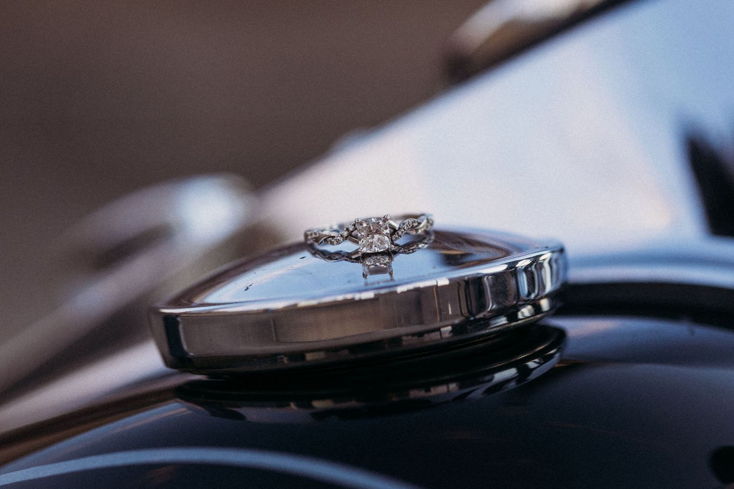 Diamond engagement ring resting on a vintage car hood ornament, showcasing elegant jewelry against classic chrome.