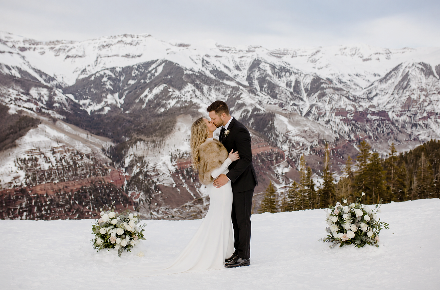 Bride and groom kissing at snowy mountain wedding ceremony with white floral arrangements and stunning alpine backdrop.