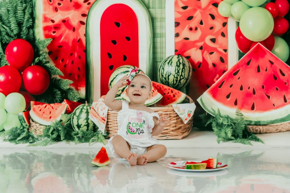 Baby girl in watermelon outfit sits smiling at cake smash photo session with festive watermelon themed backdrop.