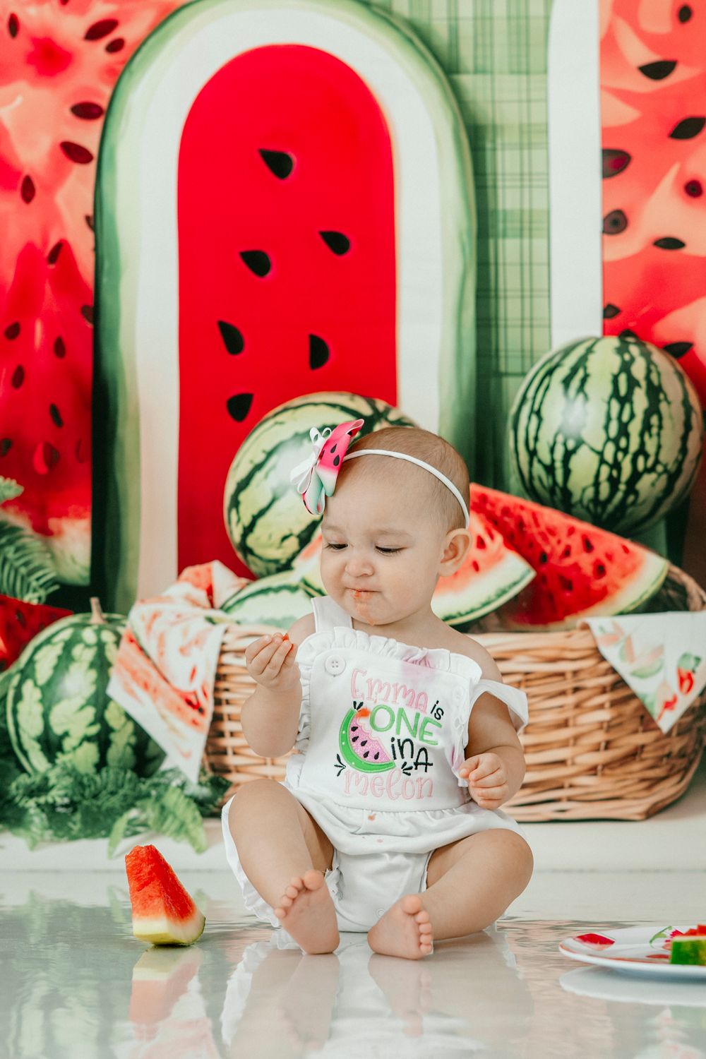 Baby girl in watermelon outfit sits on floor at one-year birthday cake smash with watermelon themed backdrop.