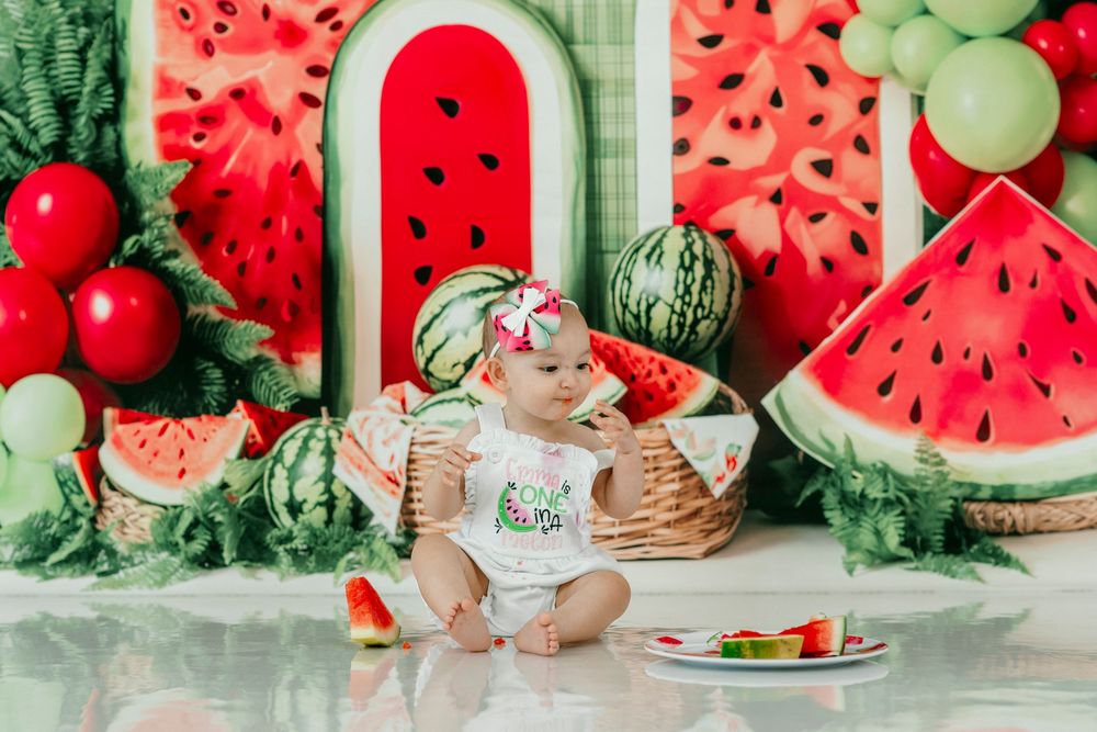 Baby girl in watermelon-themed birthday setup with decorative slices, balloons, and a smash cake plate.