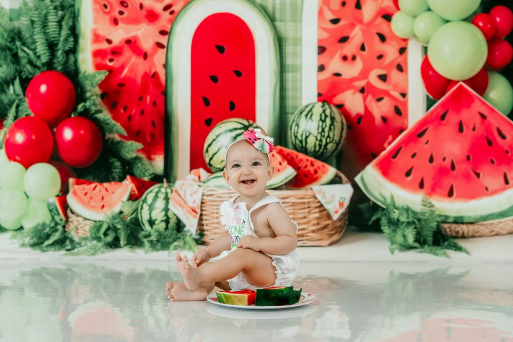 Baby girl in watermelon outfit smiles at cake smash photo session with colorful watermelon themed backdrop.