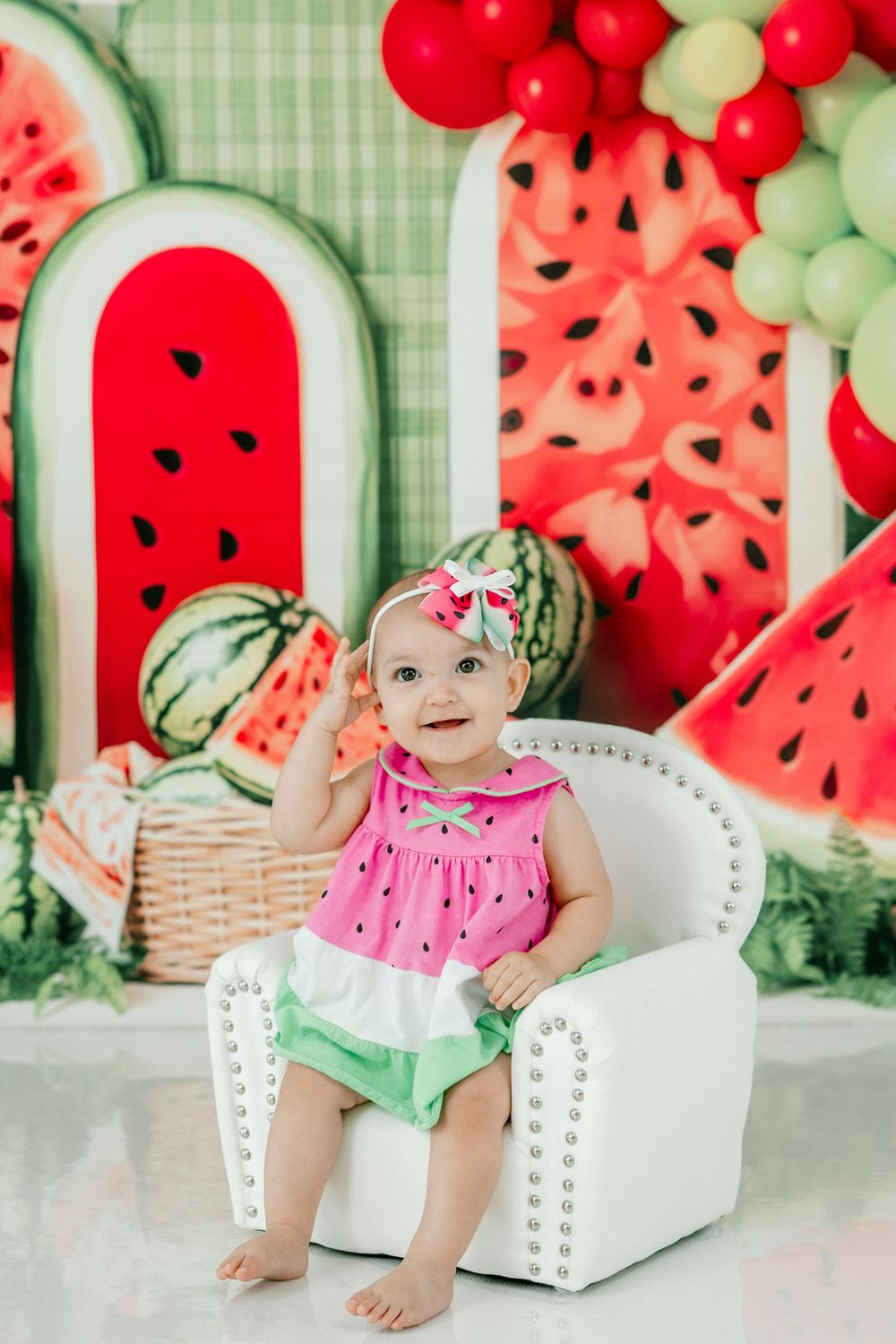 Baby girl in watermelon outfit sits smiling in white chair with festive watermelon birthday party backdrop.