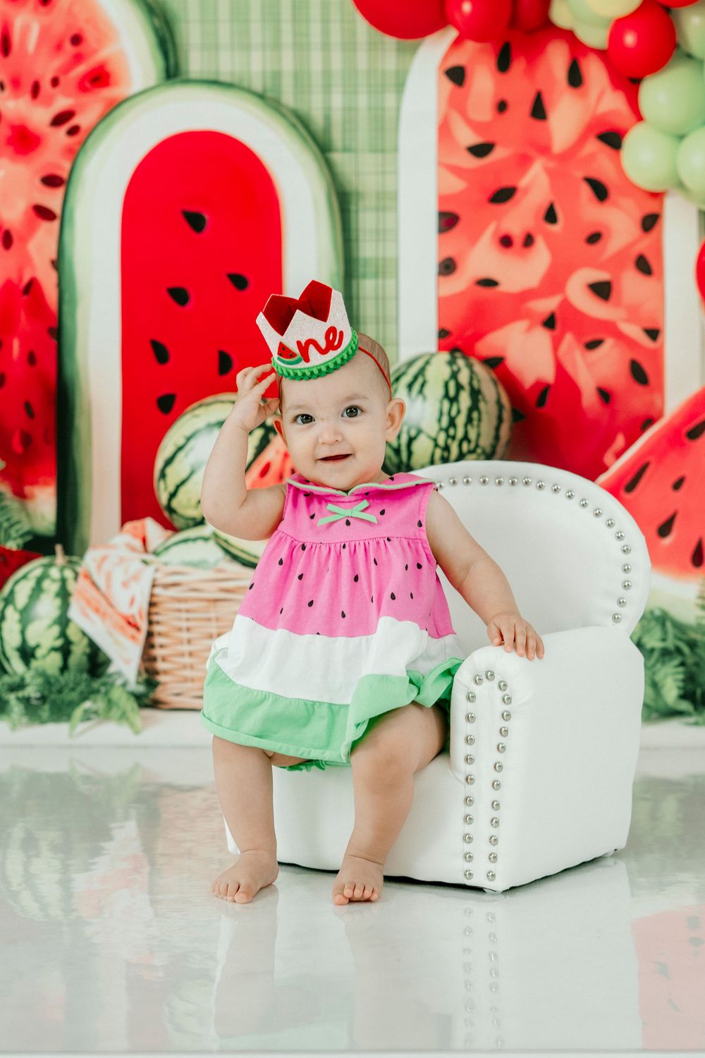 Baby girl in watermelon dress sits in white chair celebrating her first birthday with festive watermelon backdrop.
