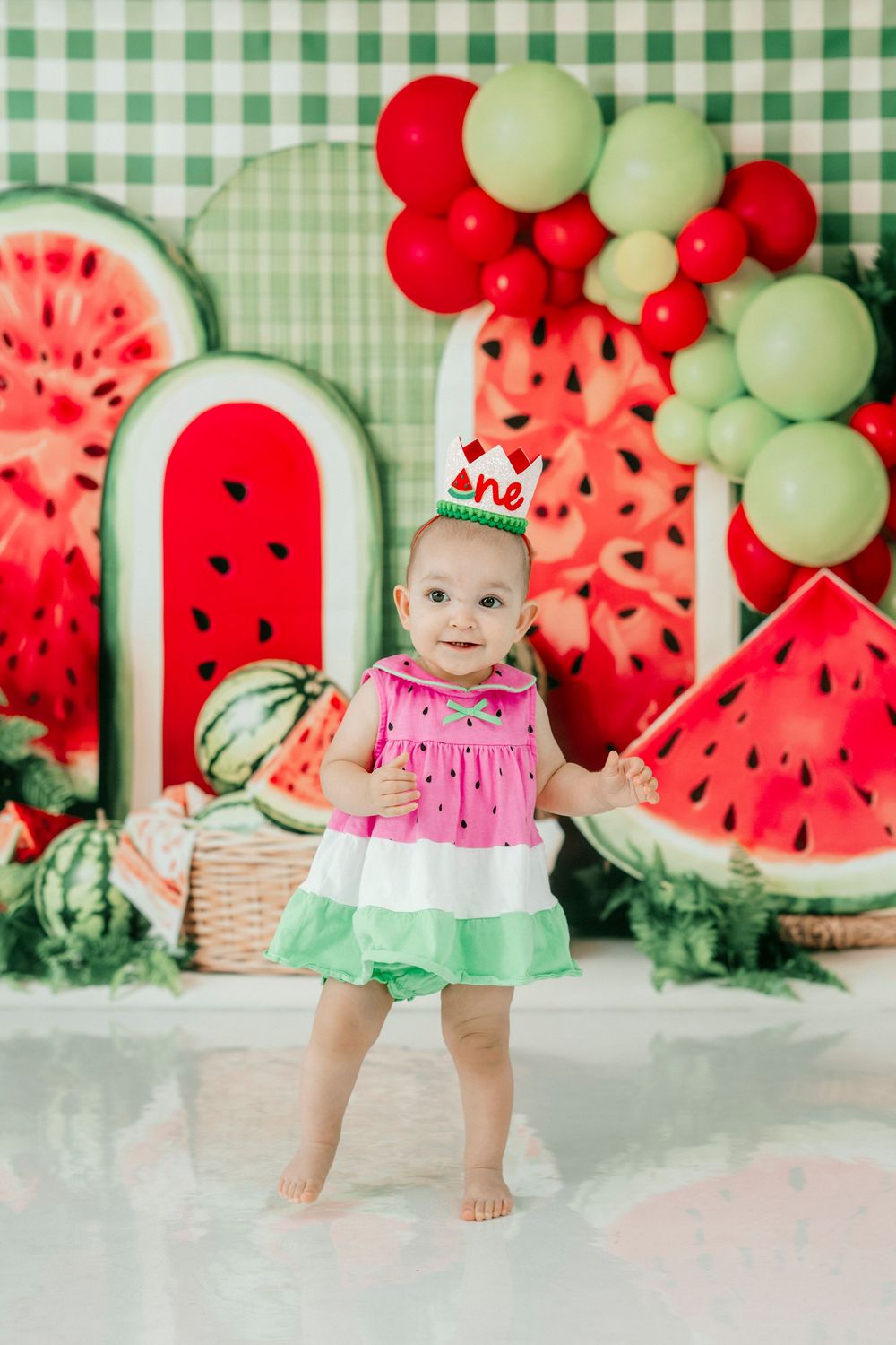 Baby girl in watermelon dress celebrates first birthday with colorful watermelon-themed backdrop and balloon decorations.