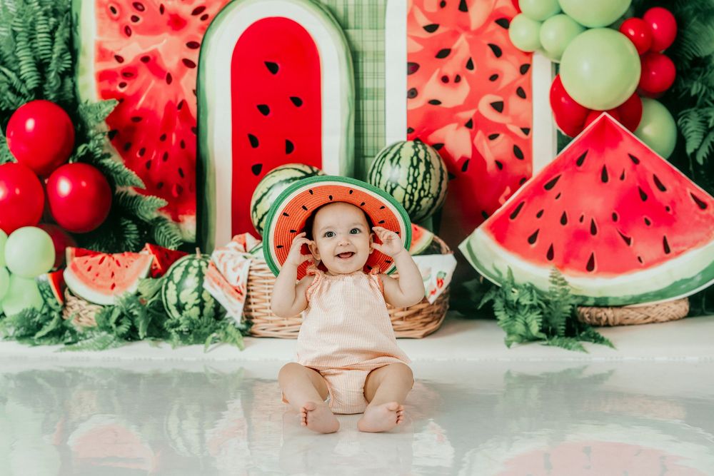 Baby girl in watermelon hat sits in basket surrounded by colorful watermelon-themed birthday photo shoot decorations.