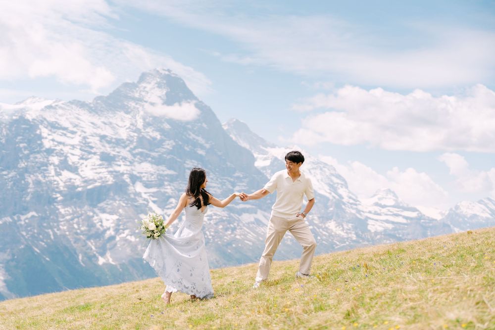 Couple holding hands on alpine meadow with snow-capped Swiss mountains backdrop, bride in white dress with bouquet.