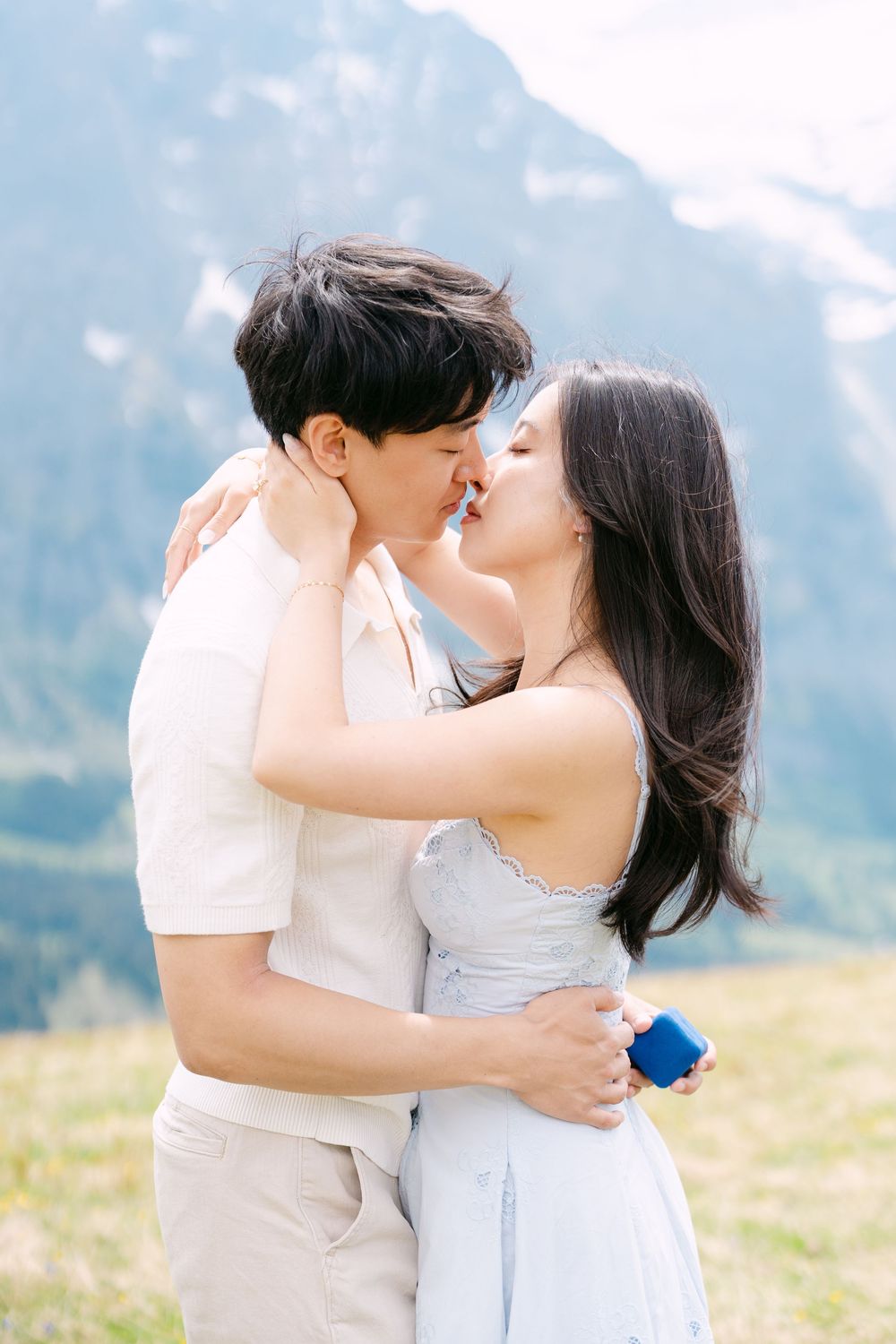 Couple sharing a romantic kiss in a mountain meadow, woman in blue dress holding a ring box during proposal.