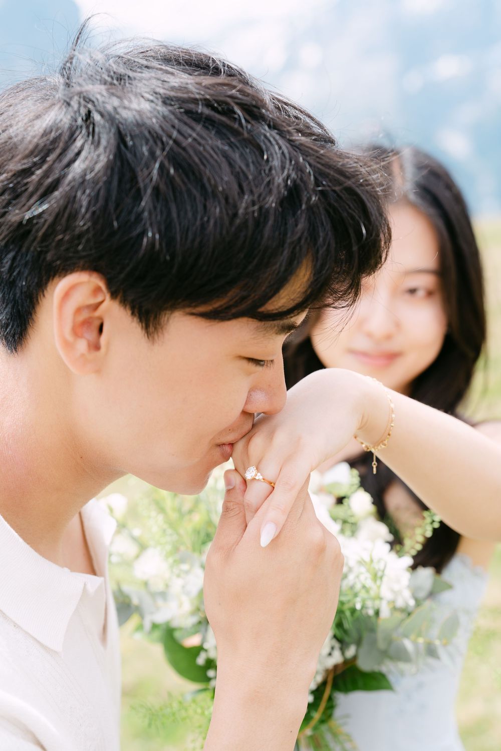 A man kisses a woman's hand as she smiles, holding white flowers outdoors in a romantic engagement moment.