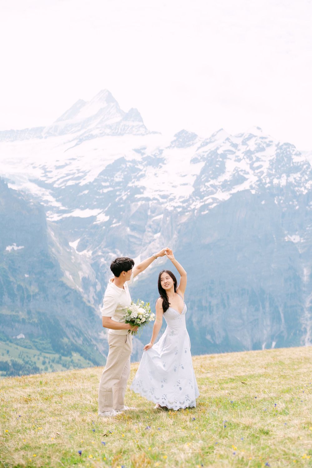 Couple dancing on alpine meadow with snow-capped Swiss mountains backdrop, bride in white dress holding bouquet.