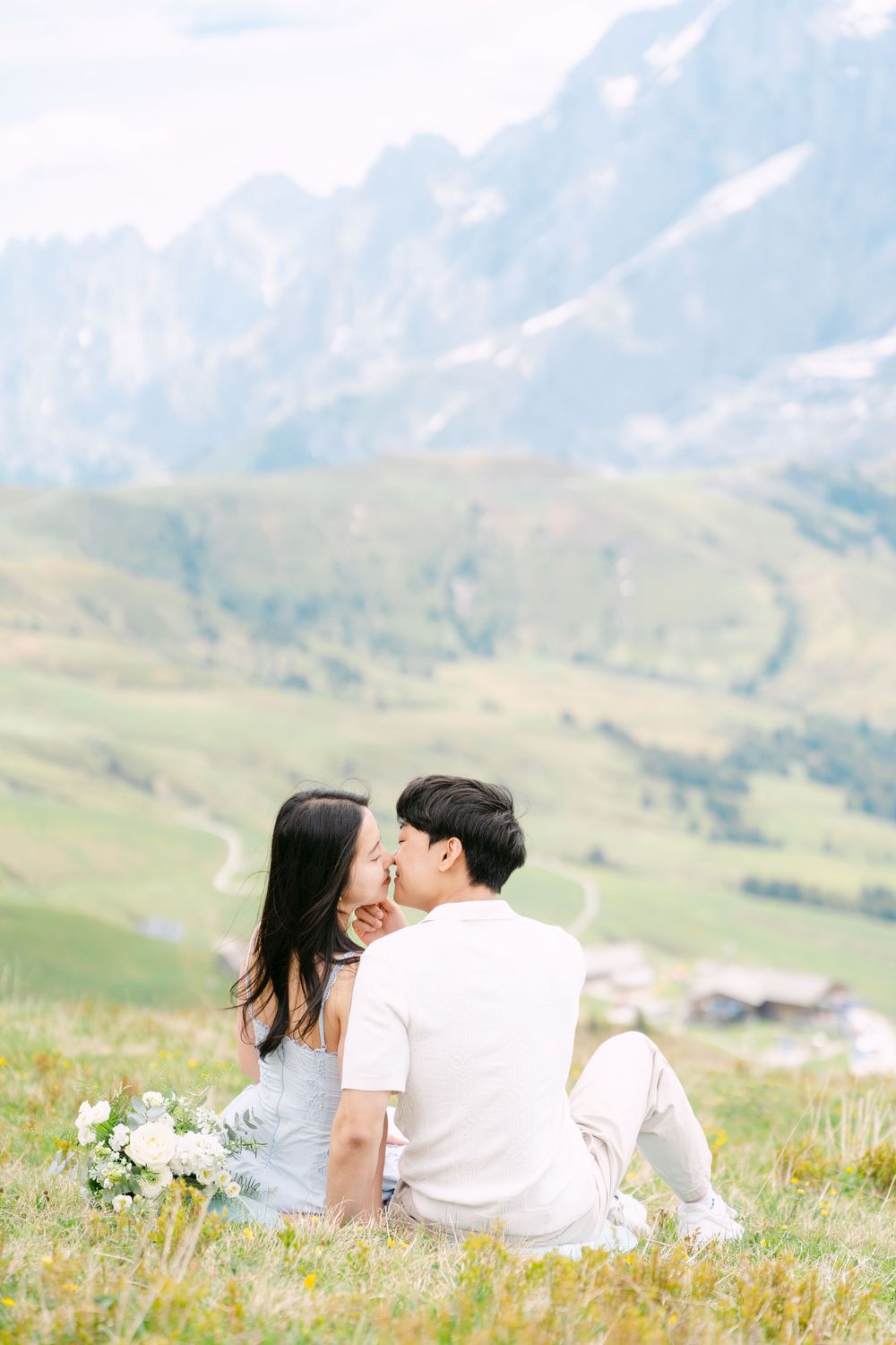 Couple sitting on alpine meadow, sharing a kiss with stunning mountain landscape backdrop, holding white bouquet.
