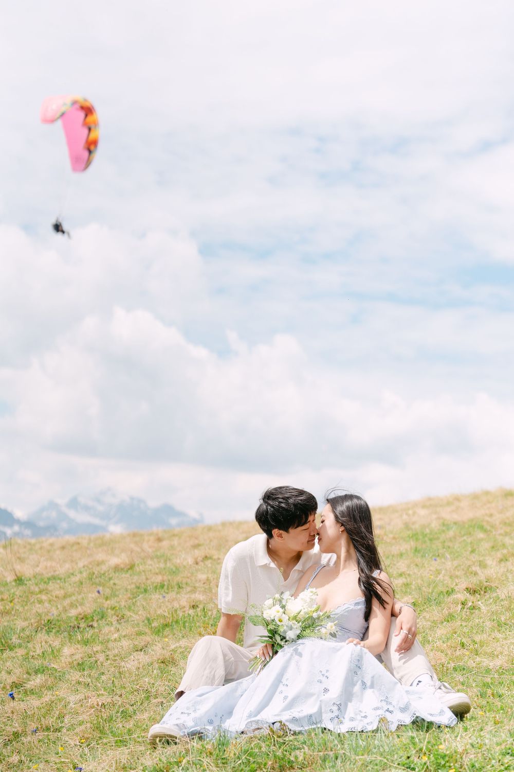 Couple sitting on grassy hillside in white outfits, sharing a romantic moment with a paraglider soaring in the cloudy sky.