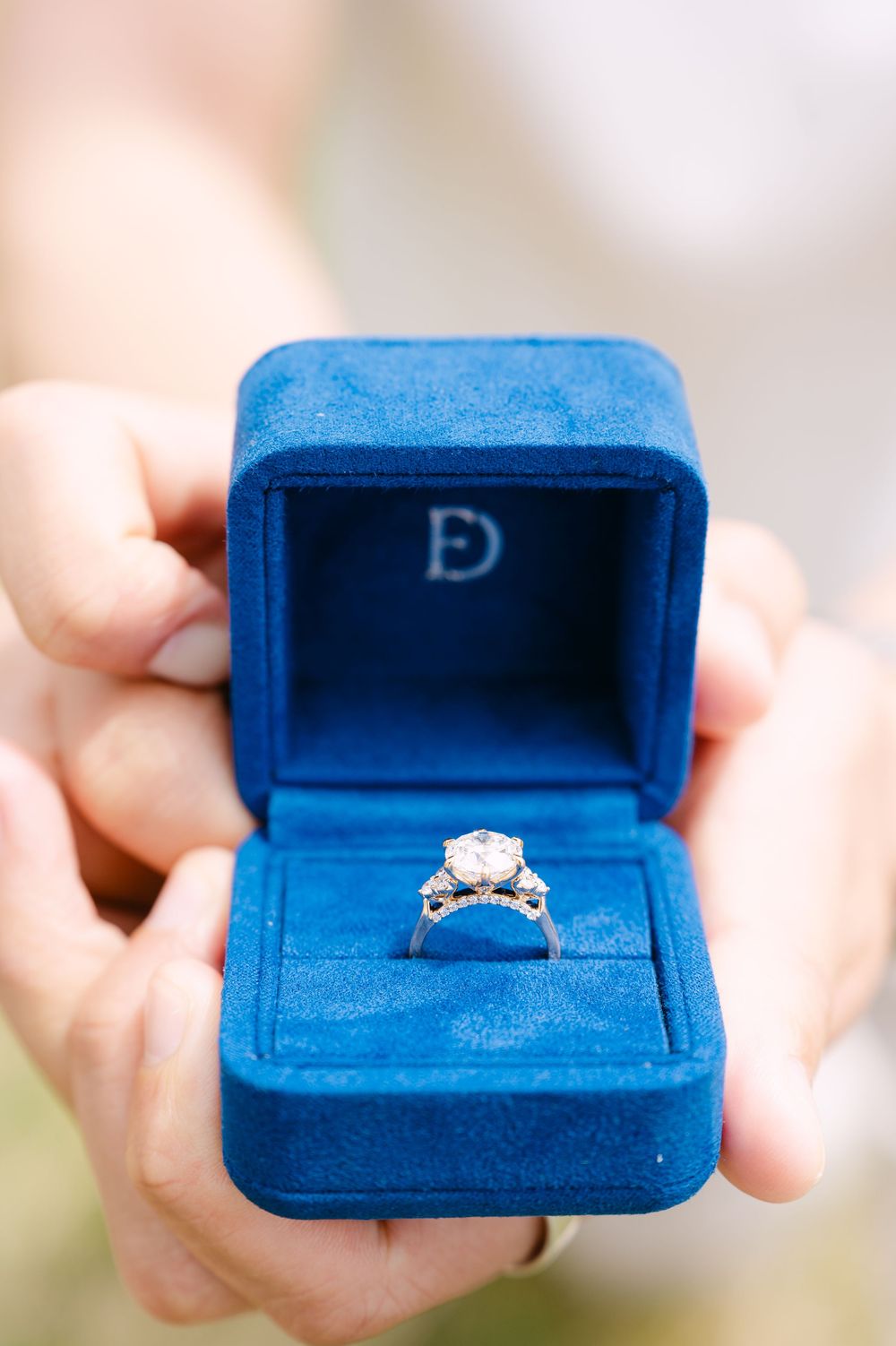 Hands holding a blue velvet ring box with a diamond solitaire engagement ring inside, monogrammed lid.