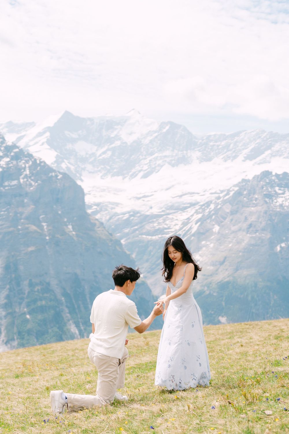 Man proposes on one knee to woman in white dress on alpine meadow with snow-capped mountains backdrop.