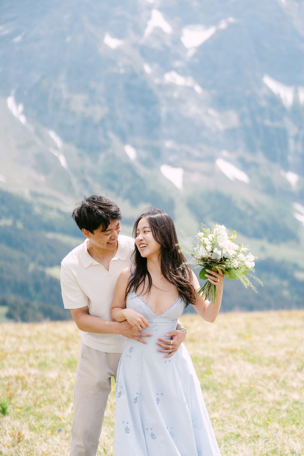 Couple embracing in alpine meadow, woman in blue dress holding white bouquet with snow-capped mountains behind them.