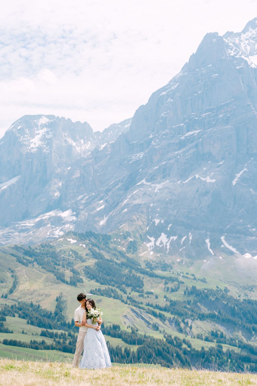 Bride and groom embracing on alpine meadow with dramatic snow-capped mountain peaks in background.