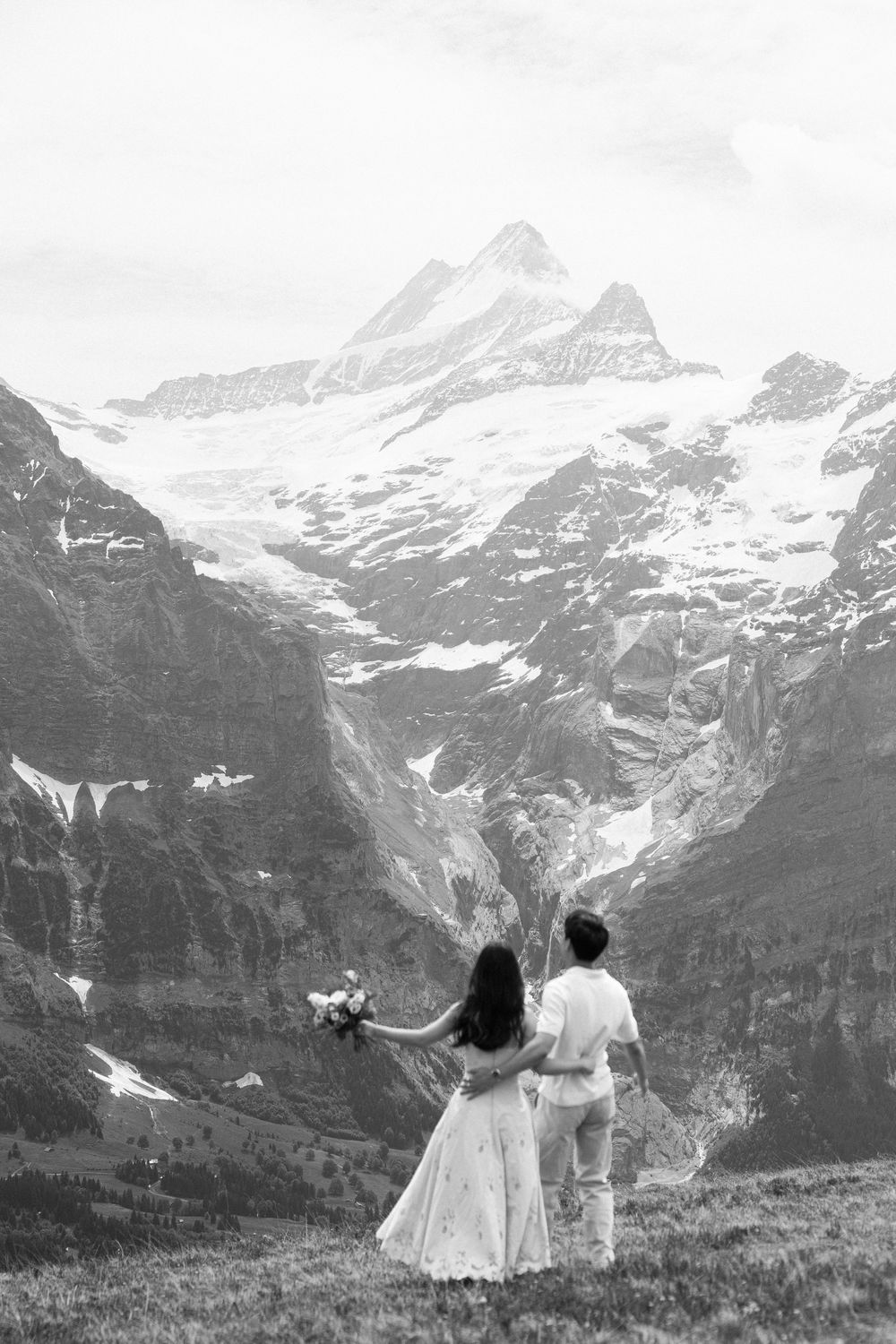 Black and white photo of a couple in wedding attire standing on a mountain overlook with snowy Alpine peaks in the background.