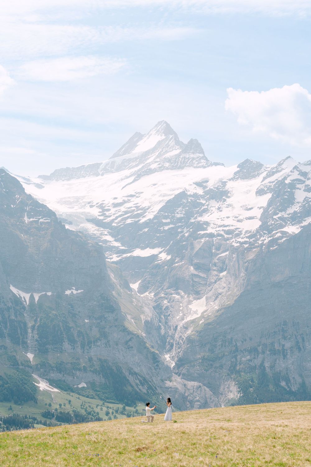 Couple standing on alpine meadow with snow-capped Swiss mountain peaks and glaciers in background under hazy sky.