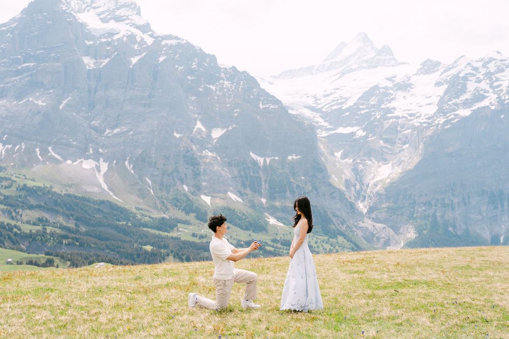 Man proposing to woman in white dress on alpine meadow with snow-capped mountains in background.