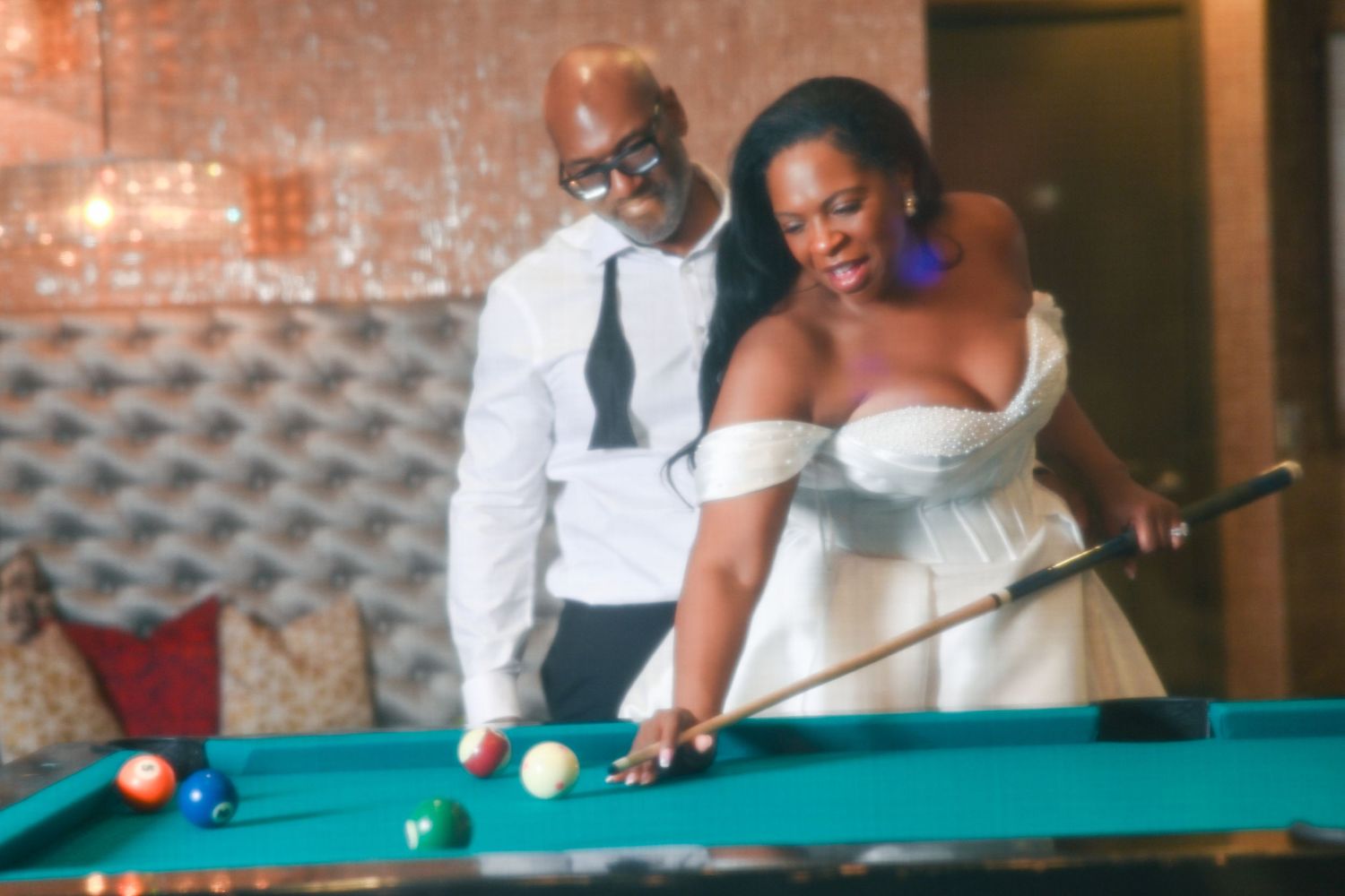 Bride in white strapless gown playing pool with groom in white shirt and tie at wedding reception.