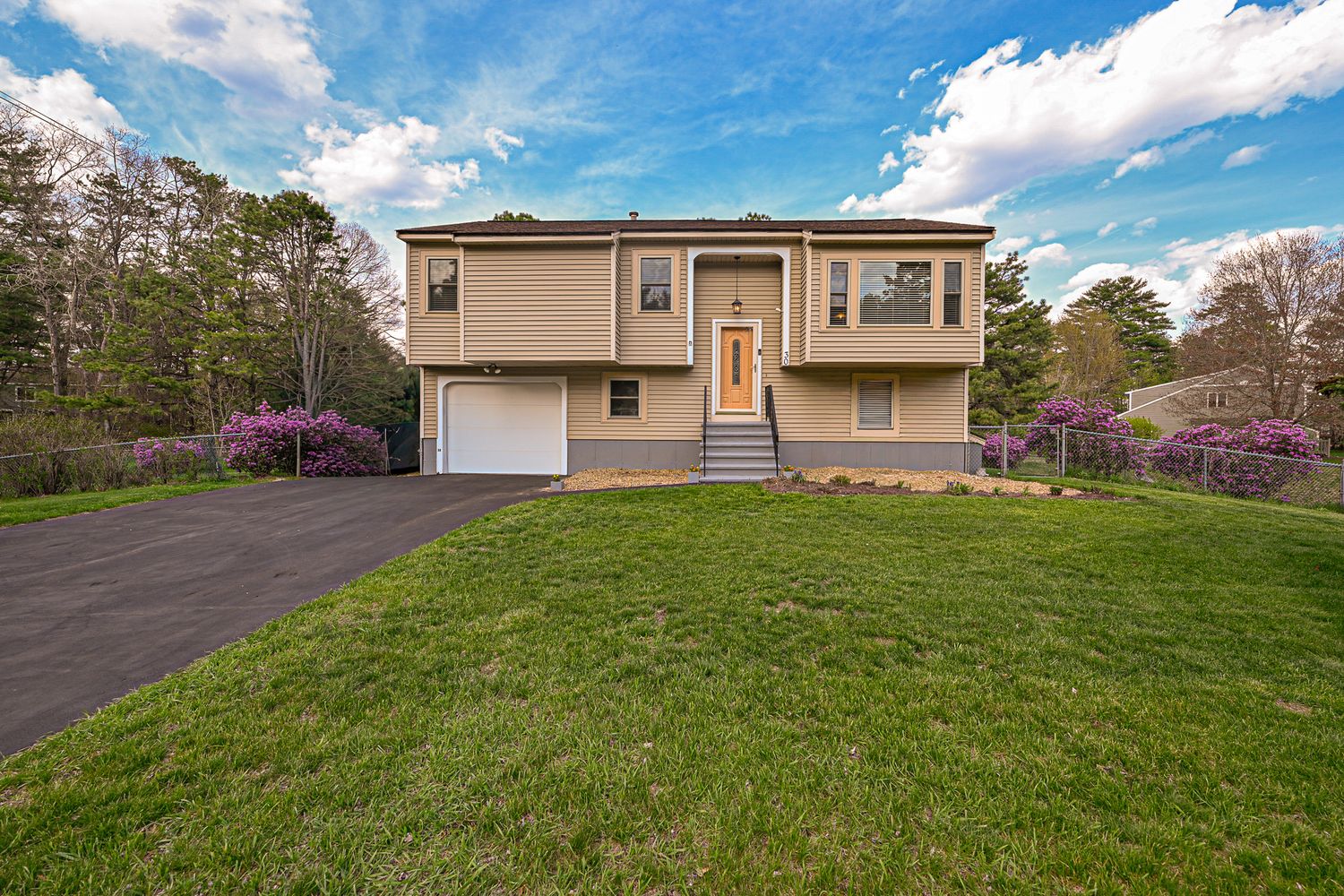 Tan split-level home with white garage, yellow front door, lush green lawn, and blooming pink trees under blue sky.