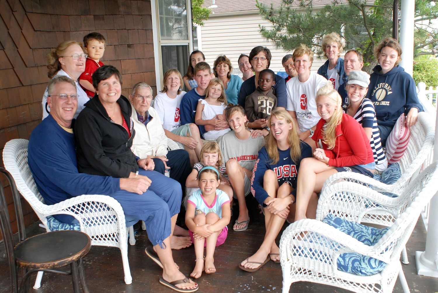 Large family group photo on a porch with white wicker furniture, smiling adults and children gathered together.